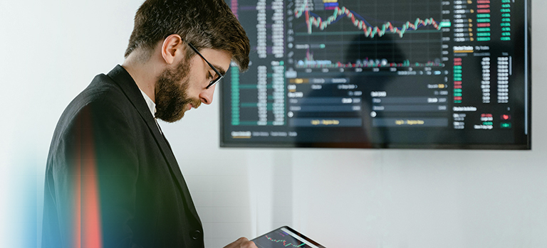 Person in business attire using a tablet in front of a large screen displaying financial charts and stock market data.