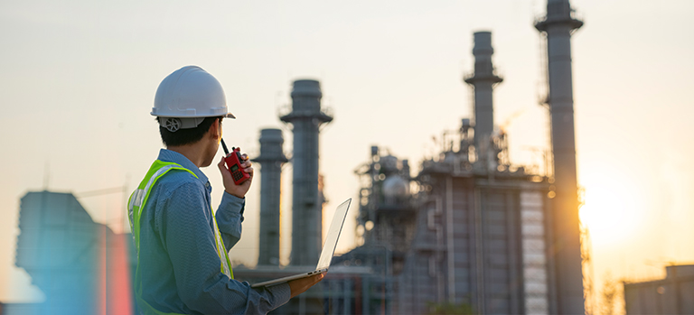 Engineer wearing a safety helmet and high-visibility vest, holding a laptop and two-way radio while observing an industrial plant with tall structures at sunset.